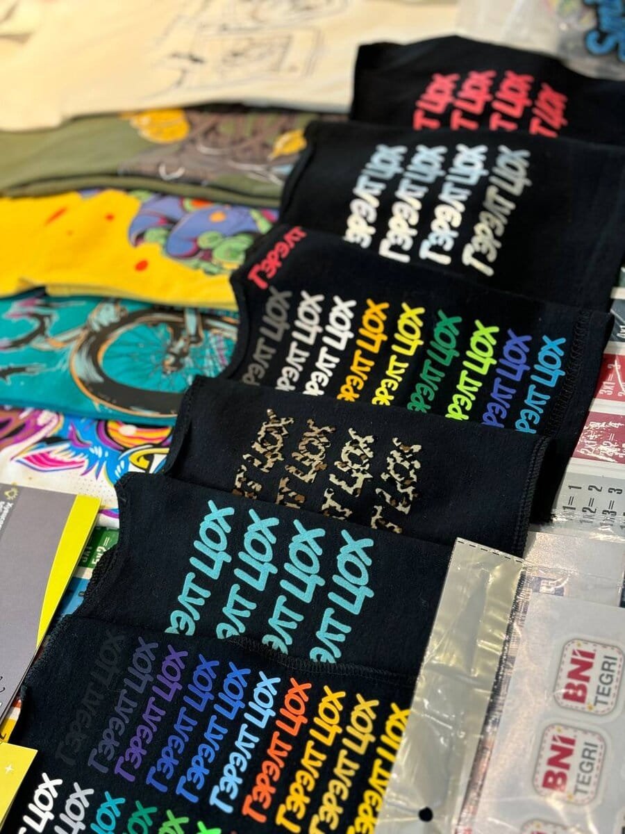 Ширээн дээр эгнүүлэн байрлуулсан, өнгөлөг, загварчлагдсан кирилл бичээс бүхий нугалсан хар подволкны дэлгэц, бусад график цамц, төрөл бүрийн эд зүйлс. - Business.MN