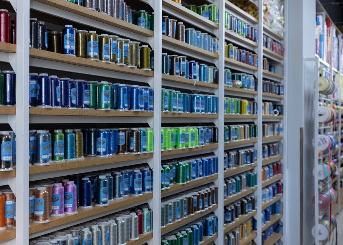 A store aisle with shelves filled with a variety of colorful drinks in cans, arranged by color. The shelves extend down the aisle, and there are visible sections of floor tiles and the ceiling.