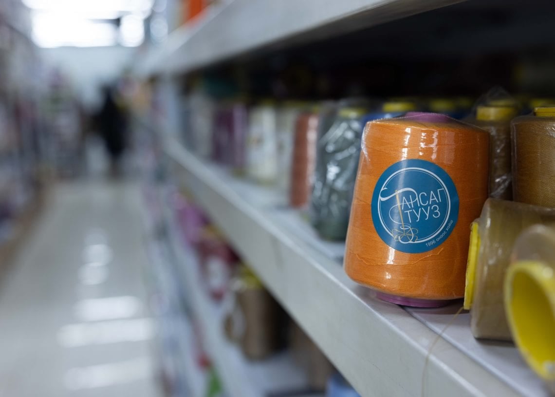 Close-up of an orange spool of thread with a blue label on a store shelf, surrounded by other colorful spools. The background shows a blurred aisle with a person walking in the distance.