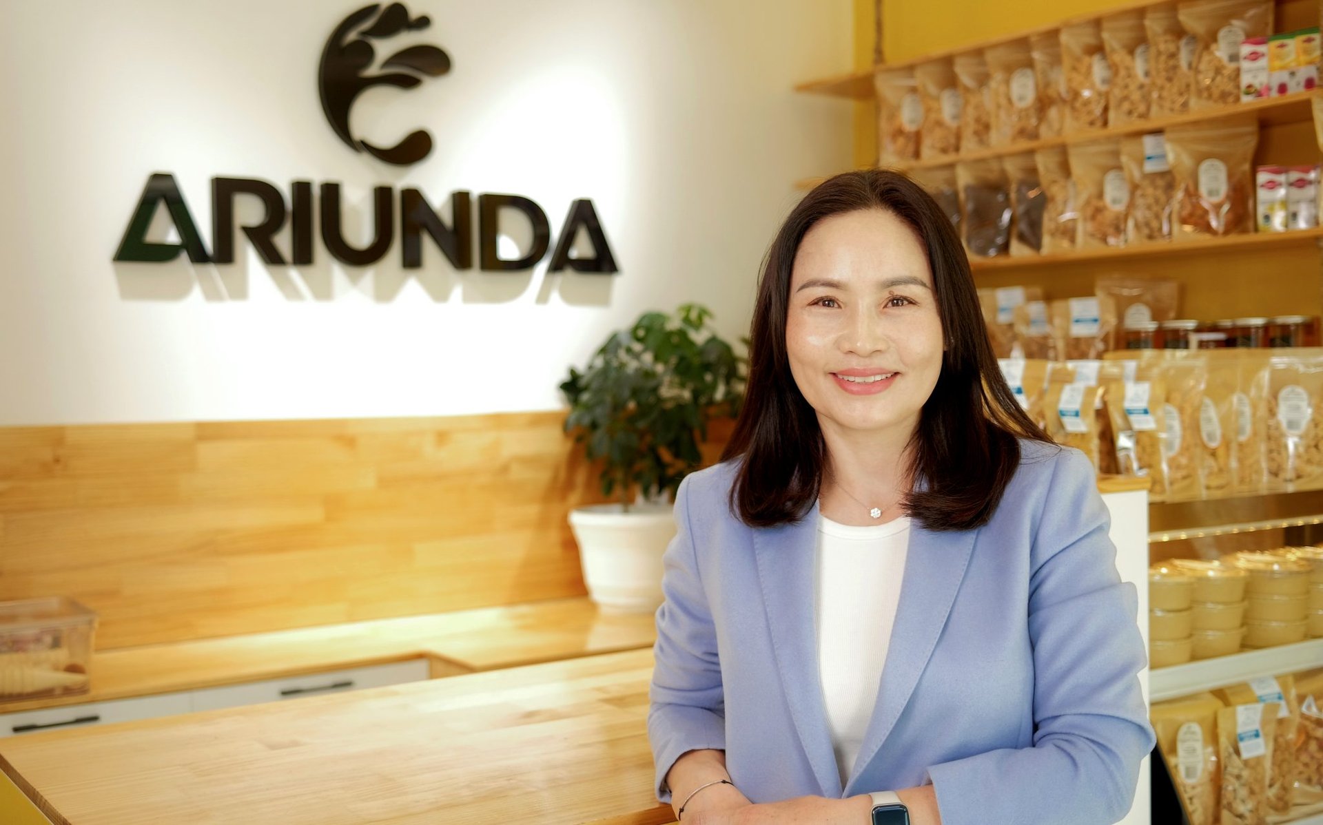 A woman in a light blue blazer stands smiling at a wooden counter in a bright shop with shelves of packaged goods. A sign behind her reads "ARIUNDA." A potted plant is visible in the background.