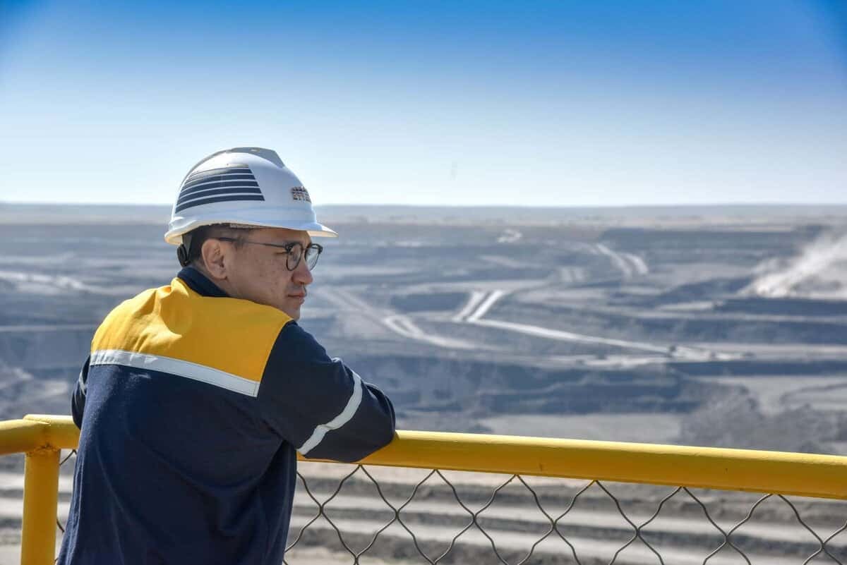 Хамгаалалтын хэрэгсэлтэй хүн цэлмэг хөх тэнгэрийн дор урд талын хашлагатай том ил уурхай руу харж байна. Энэ үзэгдэл нь нарийн хяналт, стратеги төлөвлөлт чухал байдаг бизнесийн мөн чанарыг илэрхийлдэг. - Business.MN