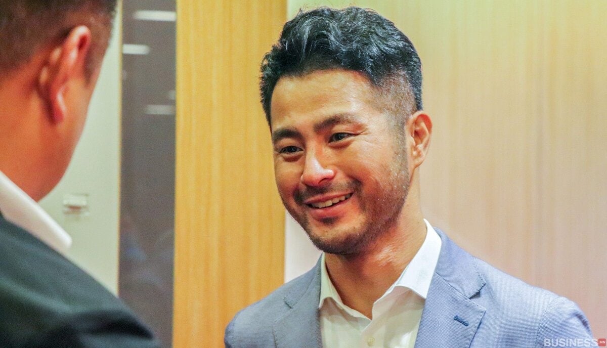 Цэнхэр костюмтай эр инээж, камерын гадуур хэн нэгэнтэй ярьж байна. Тэдгээр нь доторх бөгөөд цаана нь модон хана харагдана. - Business.MN