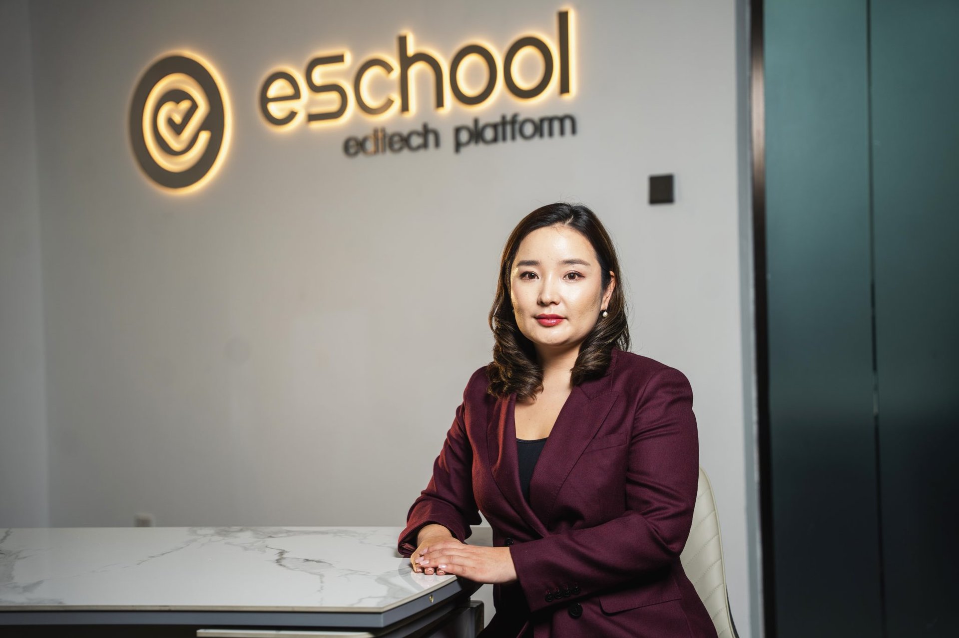 A woman in a burgundy blazer sits at a marble desk with her hands folded. Behind her, a wall features the illuminated logo of "eSchool edtech platform" in yellow. The setting appears to be an office.