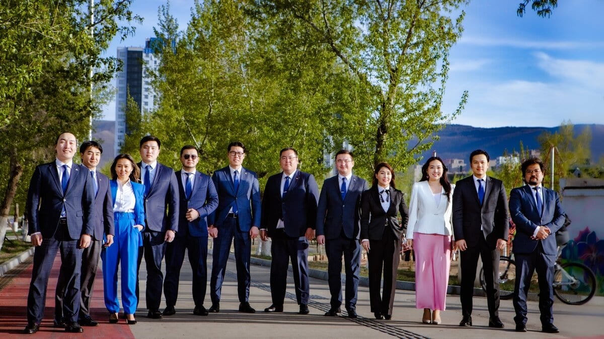 A group of twelve professionally dressed people stand in a row outdoors on a sunny day, with trees and buildings in the background. Most are wearing business suits; two women wear smart business attire with colorful skirts and pants.