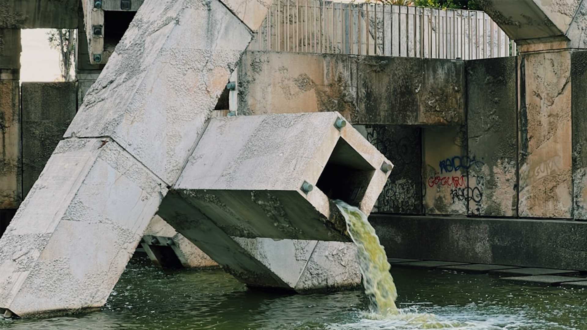A large, angular concrete structure with water flowing from a rectangular opening into a pool below, surrounded by graffiti-covered walls and metal railings.