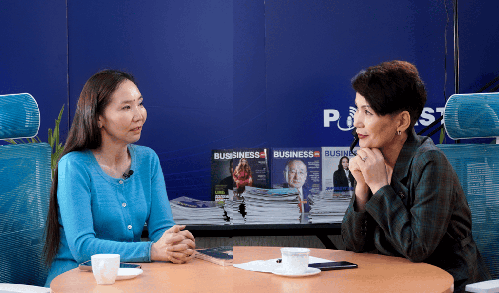 Хоёр эмэгтэй дугуй ширээний ард суугаад ярилцаж байна. Ширээн дээр кофены аяга, ухаалаг утас байна. Цаана нь цэнхэр дэвсгэр дээр "BUSINESS" гэсэн гарчигтай овоо сэтгүүл харагдаж байгаа нь "PODCAST" гэсэн бичигтэй хэсэгчлэн харагдаж байгаа нь бизнес болон энтрепренерийн сэдвүүдэд анхаарлаа хандуулж байгааг илтгэнэ. - Business.MN