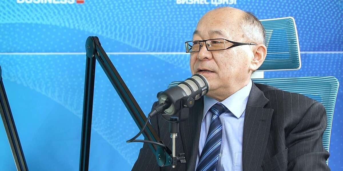 Костюм, нүдний шил зүүсэн хүн микрофоноор ярьж байна. Арын дэвсгэр нь цэнхэр өнгөтэй, цагаан бичээстэй, хүн сандал дээр сууж байна. - Business.MN