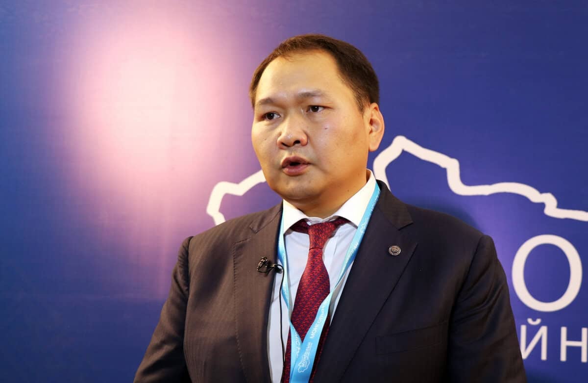 Костюм, улаан зангиа зүүсэн эр арга хэмжээн дээр ярьж байна. Түүний энгэртээ микрофон, хүзүүндээ оосортой. Арын дэвсгэр нь цэнхэр өнгөтэй, цагаан тексттэй, газрын зургийн хэсэгчилсэн тоймтой. - Business.MN