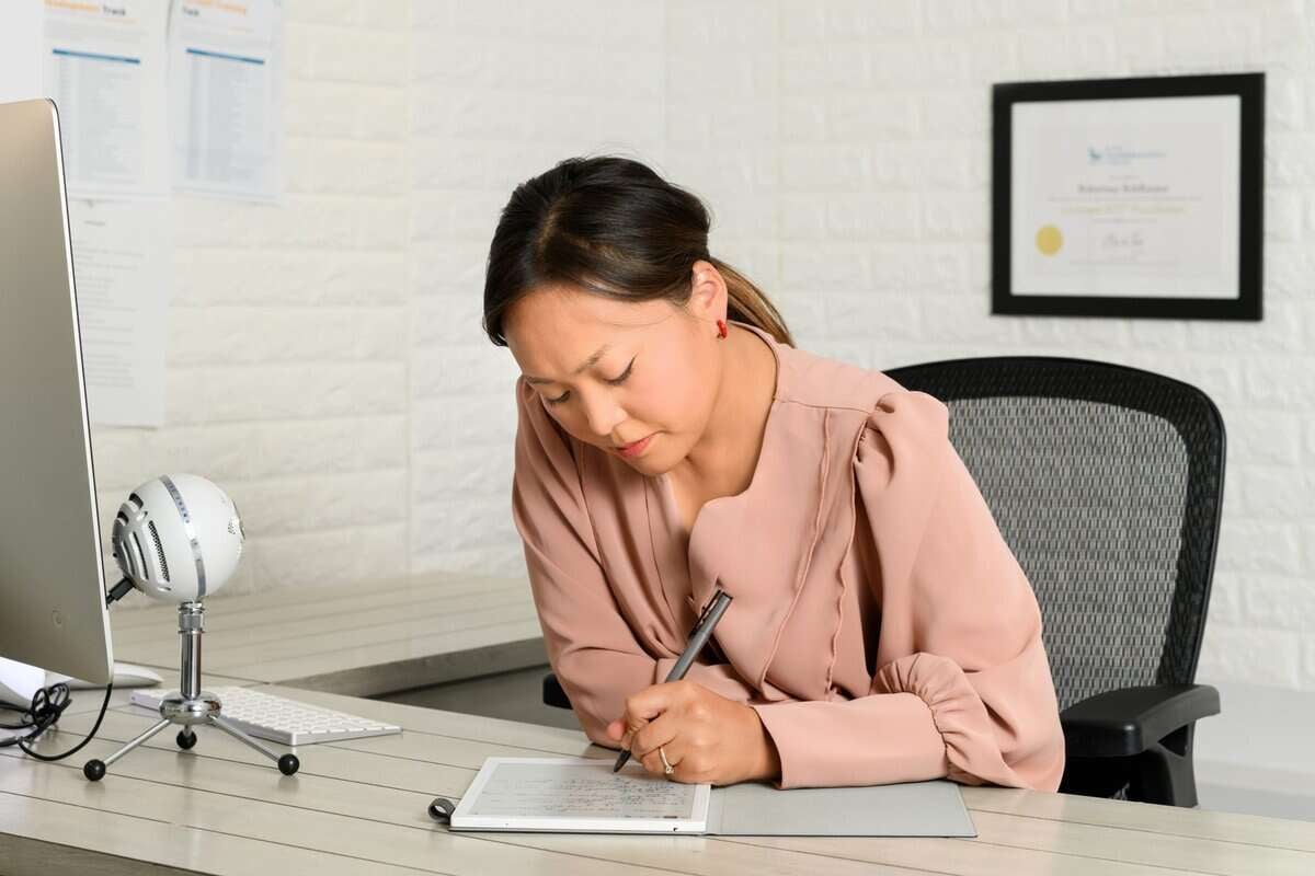 Нэг эмэгтэй албан тасалгааны ширээний ард суугаад тэмдэглэлийн дэвтэр дээр үзэг барин бичдэг. Тэр анхаарлаа төвлөрүүлж, цайвар ягаан өнгийн цамц өмссөн; the backdrop hints at Soupreneurship or зүрх сэтгэл төвтэй бизнес with a computer, microphone, and framed certificate. - Business.MN