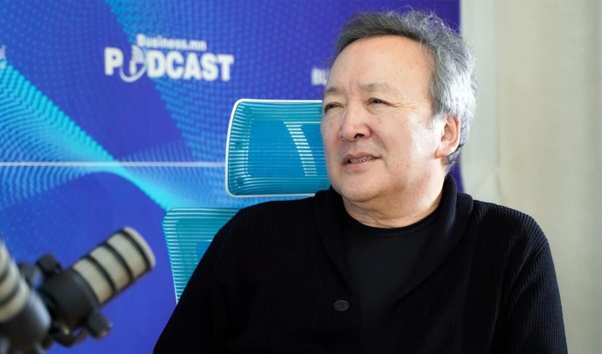 Богино саарал үстэй, хар цамц, цамц өмссөн хүн бизнесийн подкаст бичлэг хийх үеэр микрофоны өмнө сууж байна. Арын дэвсгэр дээр "Business.mn PODCAST" гэсэн бичвэр бүхий цэнхэр дэлгэц, минималист дууны долгионы хэв маягийг харуулсан нь сонирхолтой энтрепренерүүдийн хэлэлцүүлэг өрнүүлэх боломжийг бүрдүүлдэг. - Business.MN