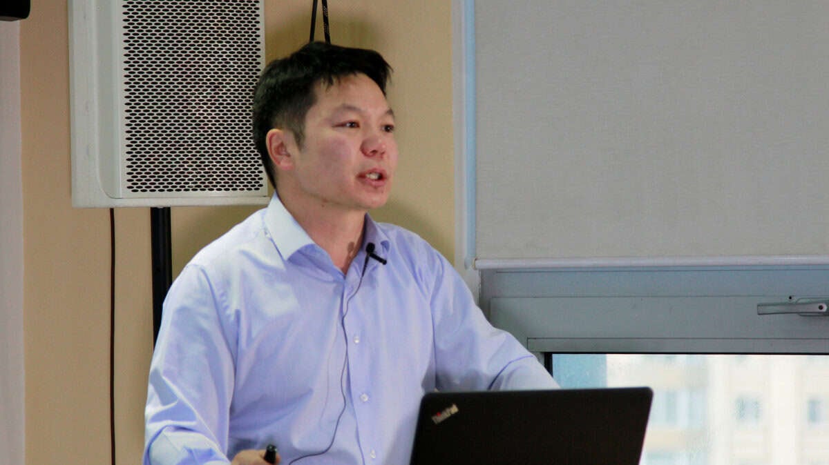 Цайвар цэнхэр цамцтай хүн илтгэл тавьж байна. Тэр товчлуур бариад зөөврийн компьютерын өмнө микрофон чихэвчтэй зогсож байна. Цаана нь том чанга яригч, цонх харагдана. - Business.MN