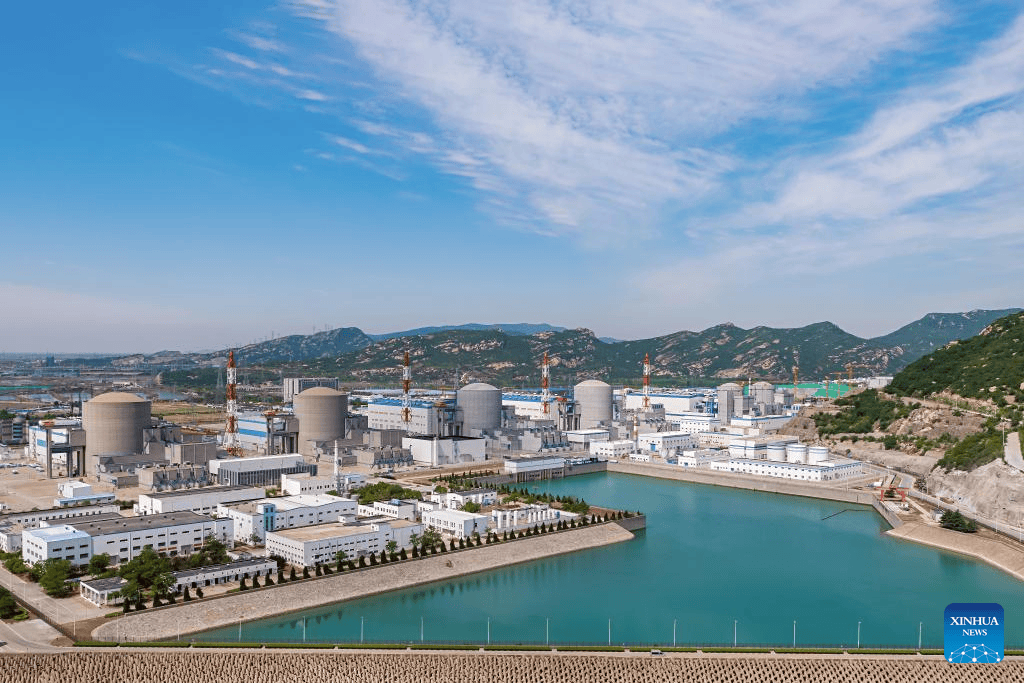 A large power plant complex with multiple cylindrical cooling towers and buildings sits beside a reservoir, surrounded by green hills under a blue sky with scattered clouds.
