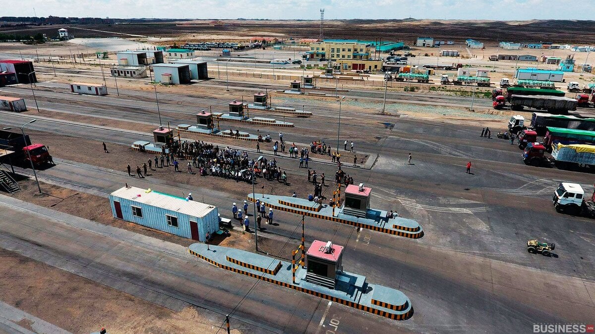 Жинлүүрийн станц, ачааны машины дэргэд цугласан хэсэг бүлэг хүмүүстэй өргөн үйлдвэрлэлийн бүсийг агаараас харах. Ландшафт нь том задгай талбай, хэд хэдэн барилга байгууламж, ар талдаа тархай бутархай машинуудыг харуулдаг. - Business.MN