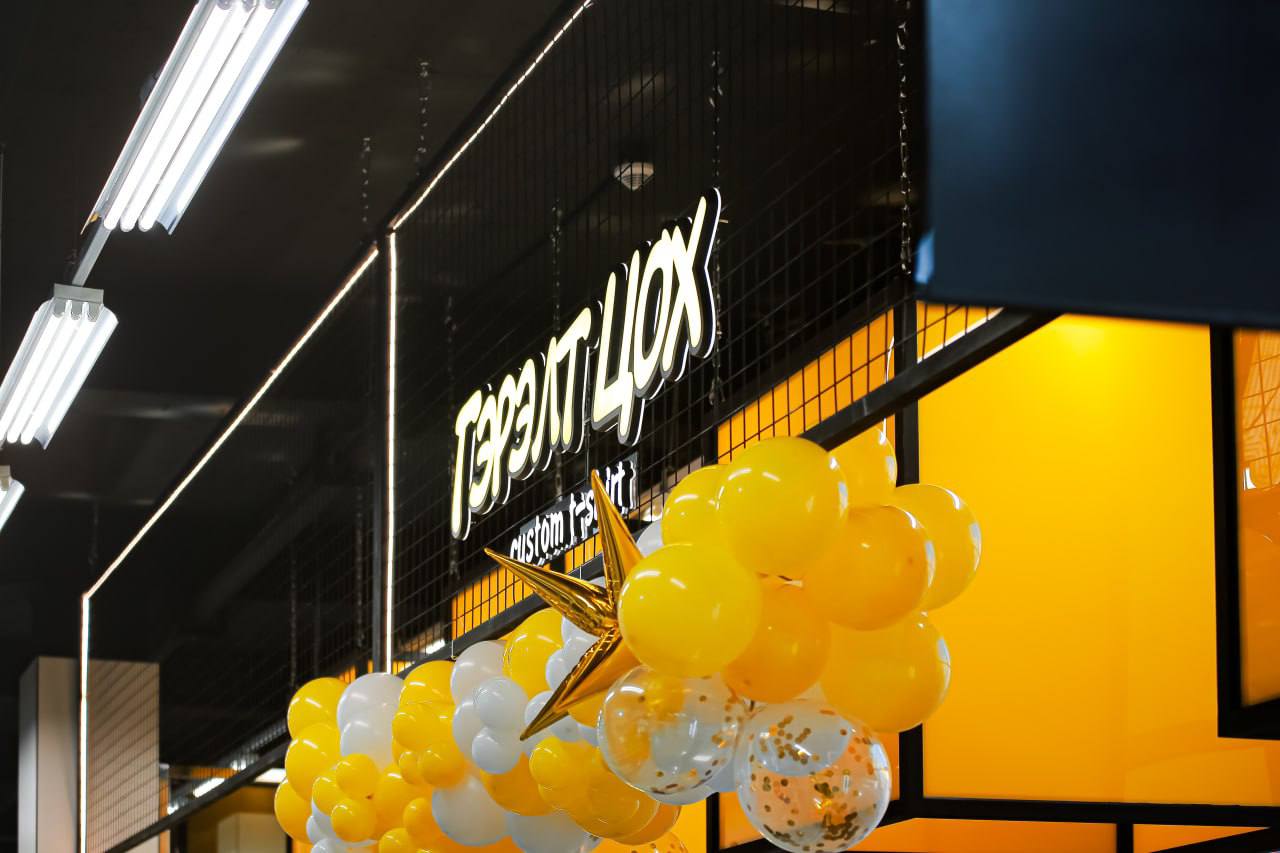 Кирилл үсэг бүхий тод шар, хар тэмдэг бүхий дэлгүүрийн лангуу. Цагаан, шар, алтлаг өнгийн бөмбөлөгүүд дэлгүүрийн нүүрийг чимдэг. Гэрэлтүүлэг нь орчин үеийн, флюресцент юм. - Business.MN