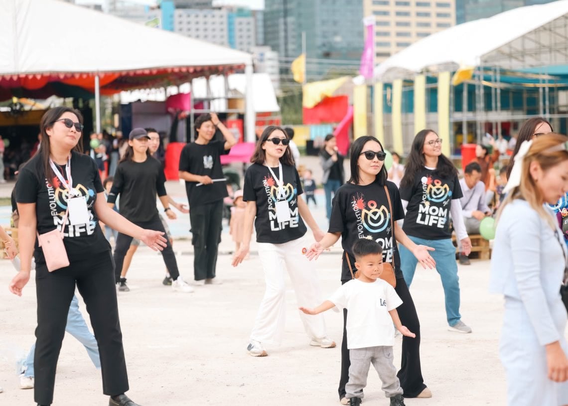A group of people in casual attire participates in a dance activity outdoors. They are wearing matching black shirts, and a young child stands among them. Colorful tents and a cityscape are visible in the background.