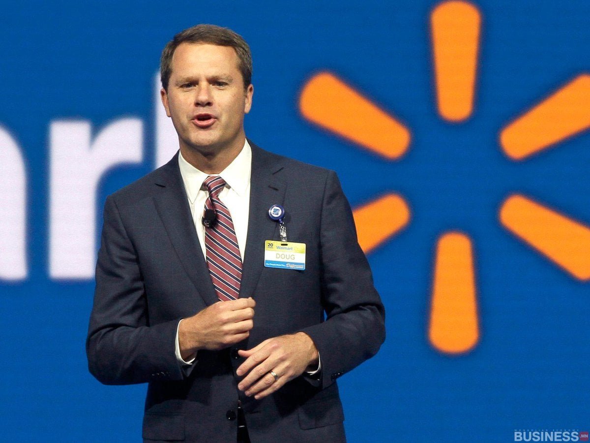 Костюм, судалтай зангиа зүүсэн эр тайзан дээр зогсож, нэрийн тэмдэг харагдана. Арын дэвсгэр нь хэсэгчилсэн лого, цэнхэр дэвсгэрийг харуулж байна. Тэрээр ярихдаа гараараа дохио зангаагаар сүй тавьсан бололтой. - Business.MN