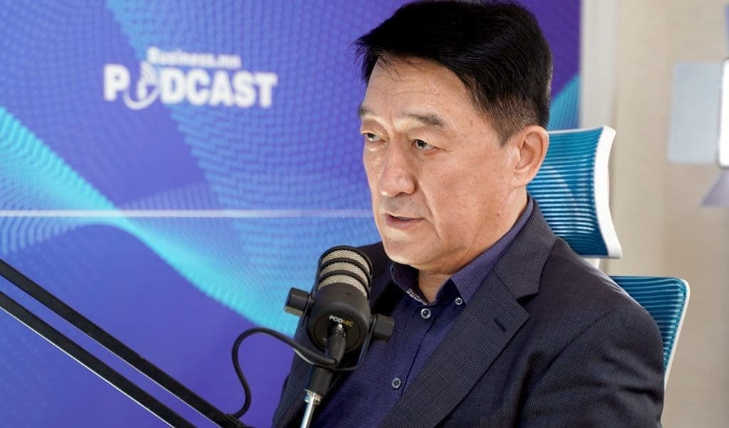 Нэг хүн сандал дээр суугаад микрофоноор ярьж байна. Арын дэвсгэр дээр "PODCAST" гэсэн үг бүхий цэнхэр өнгийн градиент тод харагдаж байна. Тэрээр бизнес болон компанийн ойлголтын талаар бичлэг хийх эсвэл шууд нэвтрүүлэг хийдэг энтрепренер бололтой. - Business.MN