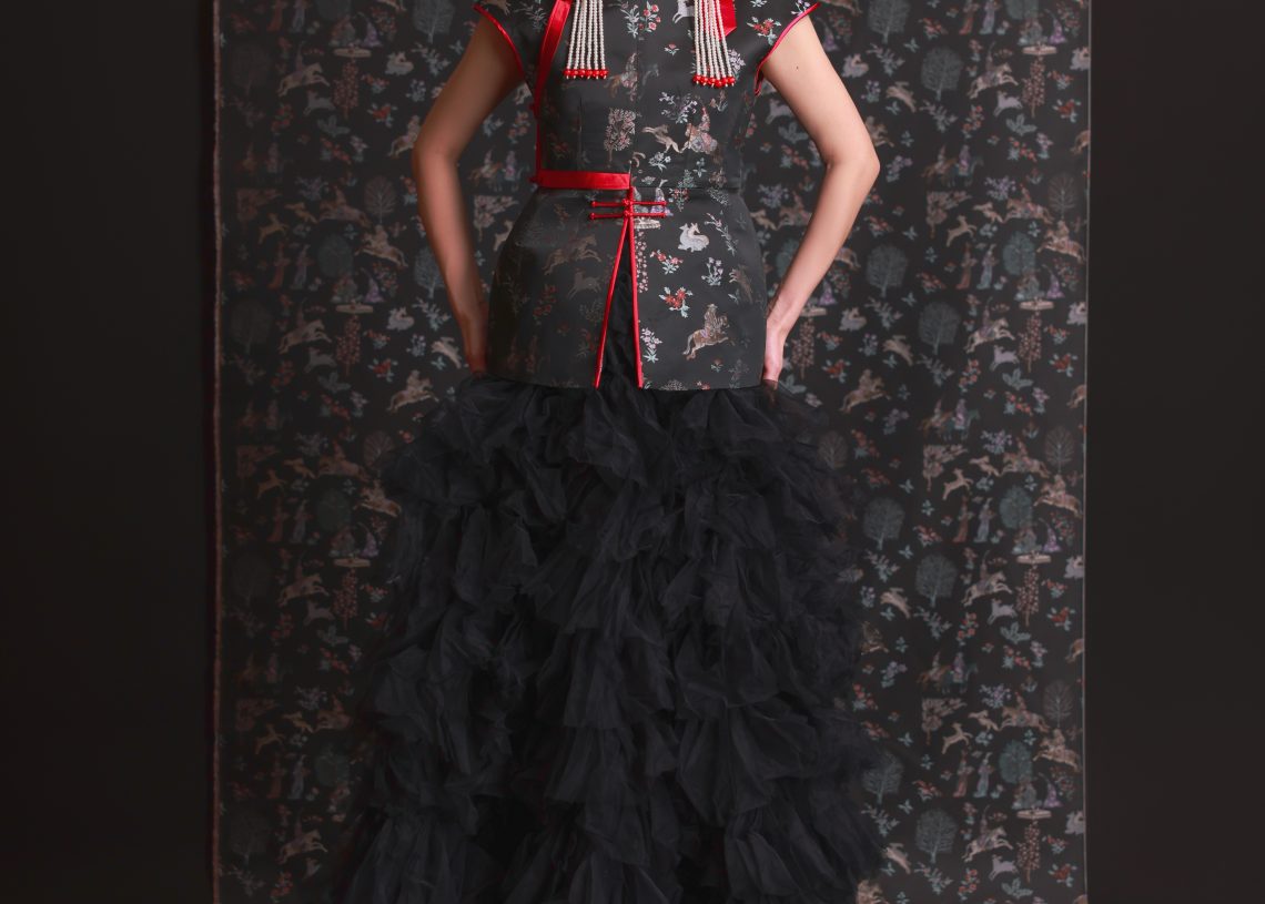 Эмэгтэй хүн харанхуй, цэцэгсийн хээтэй дэвсгэр дээр зогсож, гоёл чимэглэлийн толгойн зүүлт, улаан өнгийн өргөлттэй цэцэгсийн дээд өмд, уламжлалт болон орчин үеийн загварын элементүүдийг хослуулсан том хар tulle юбка өмссөн байна. - Business.MN