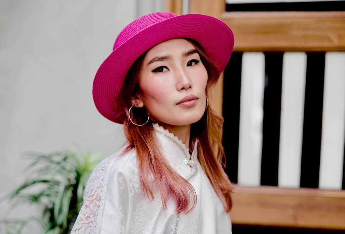 Тод ягаан малгай, том цагираг ээмэг зүүсэн, ягаан өнгийн урт үстэй хүн. Тэд ноцтой, бизнес эрхэлдэг илэрхийлэлтэй бөгөөд цагаан нэхсэн тор өмсөж, модон байгууламж, ногоон байгууламжийн өмнө зогсож байна. - Business.MN