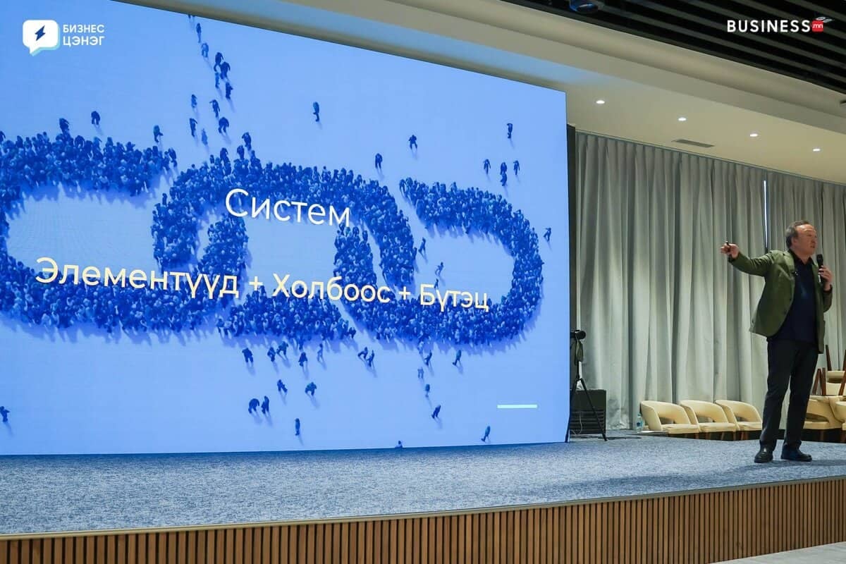 A speaker stands on a stage next to a large screen displaying a Russian presentation slide with abstract blue figures. The background shows a modern conference room with chairs.