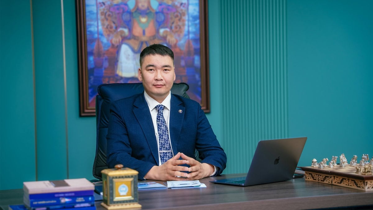 A man in a blue suit sits at a desk with a laptop, papers, books, and a chess set, symbolizing how сайн хуулийн зөвлөгөө таны бизнесийн ирээдүйг заах луужин болохыг харуулна. A framed artwork hangs on the teal-colored wall behind him.