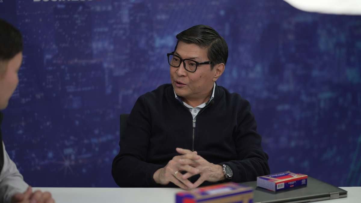 Нүдний шил, бараан ноосон цамц өмссөн эрэгтэй ширээний ард суугаад царай нь хэсэгчлэн харагдах өөр хүнтэй ярилцаж байна. Арын дэвсгэр нь цэнхэр өнгийн хотын өнгө төрхийг харуулсан бөгөөд энэ нь тухайн газрын хөл хөдөлгөөн ихтэй бизнесийг илэрхийлдэг. Эрэгтэй хүний өмнө ширээн дээр хоёр тэгш өнцөгт хайрцаг тавьдаг. - Business.MN