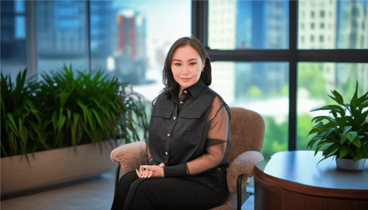 A person dressed in a black outfit sits on a chair in a modern office. Large windows reveal the city's flamme of lights, while indoor plants flank the scene. The atmosphere is calm and professional.
