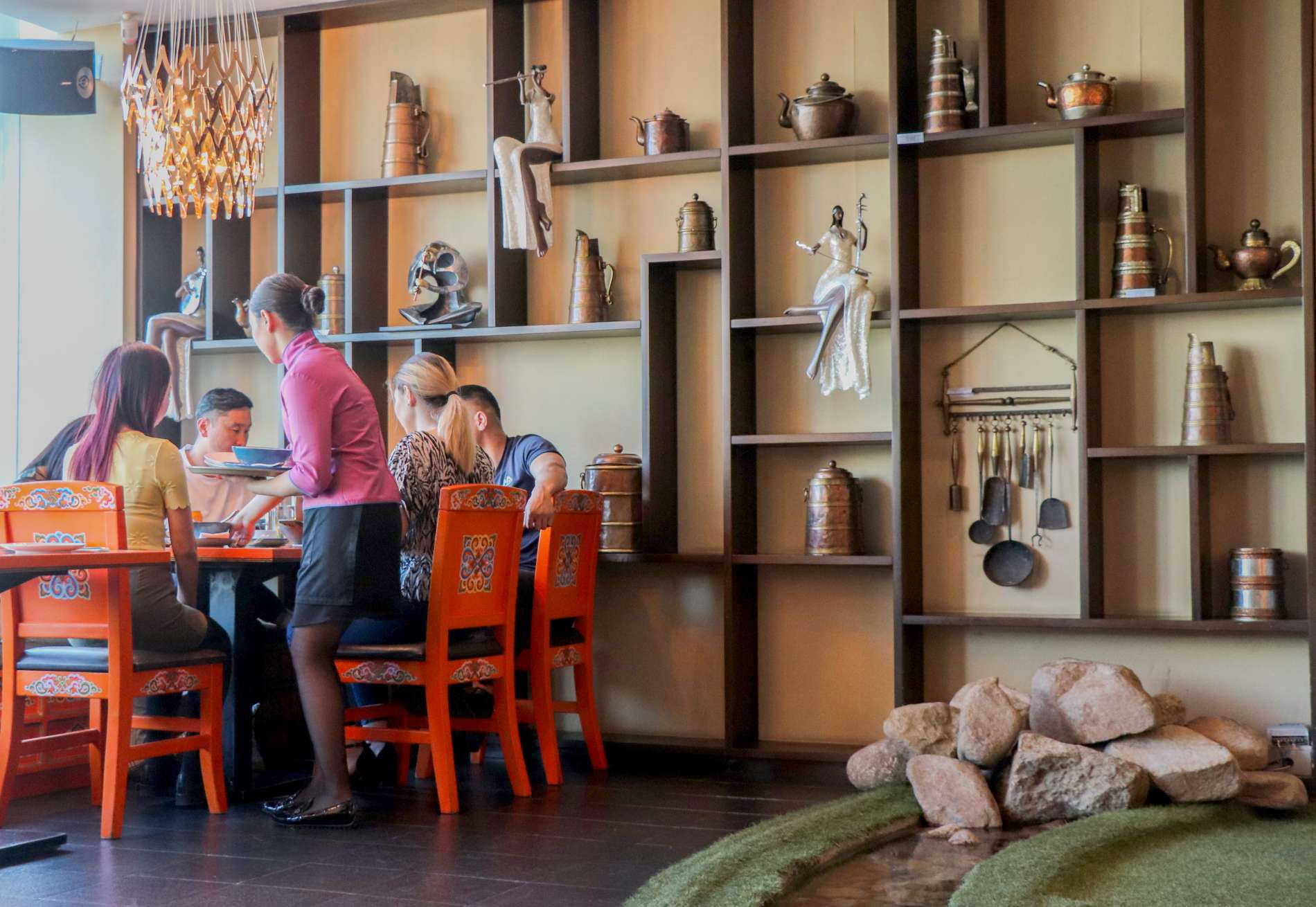 Уламжлалт вааран эдлэл, баримал бүхий нарийн төвөгтэй ханын дэлгэц бүхий рестораны ширээний ард сууж буй хэсэг хүмүүс. Ширээний дэргэд сервер зогсож байна. Чимэглэл нь урд талын хадны онцлог, дээр нь гоёл чимэглэлийн гэрэлтүүлгийг агуулдаг. - Business.MN