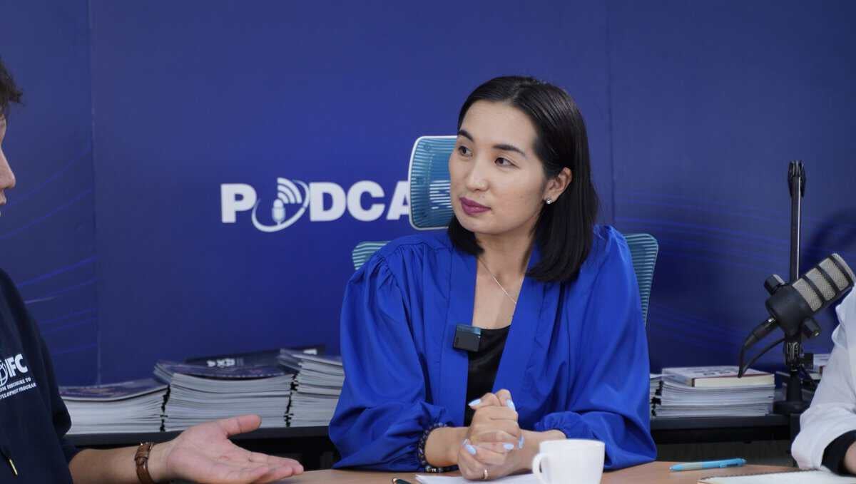 Цэнхэр цамц өмссөн эмэгтэй хүзүүвчдээ микрофон зүүсэн ширээний ард сууж байна. Тэр камерын гадуур хэн нэгэнтэй ярилцаж байгаа энтрепренер бололтой. Арын дэвсгэр дээр хэсэгчлэн харагдах "PODCAST" гэсэн бичигтэй цэнхэр туг байна. Ширээн дээр овоо бизнесийн ном байна. - Business.MN