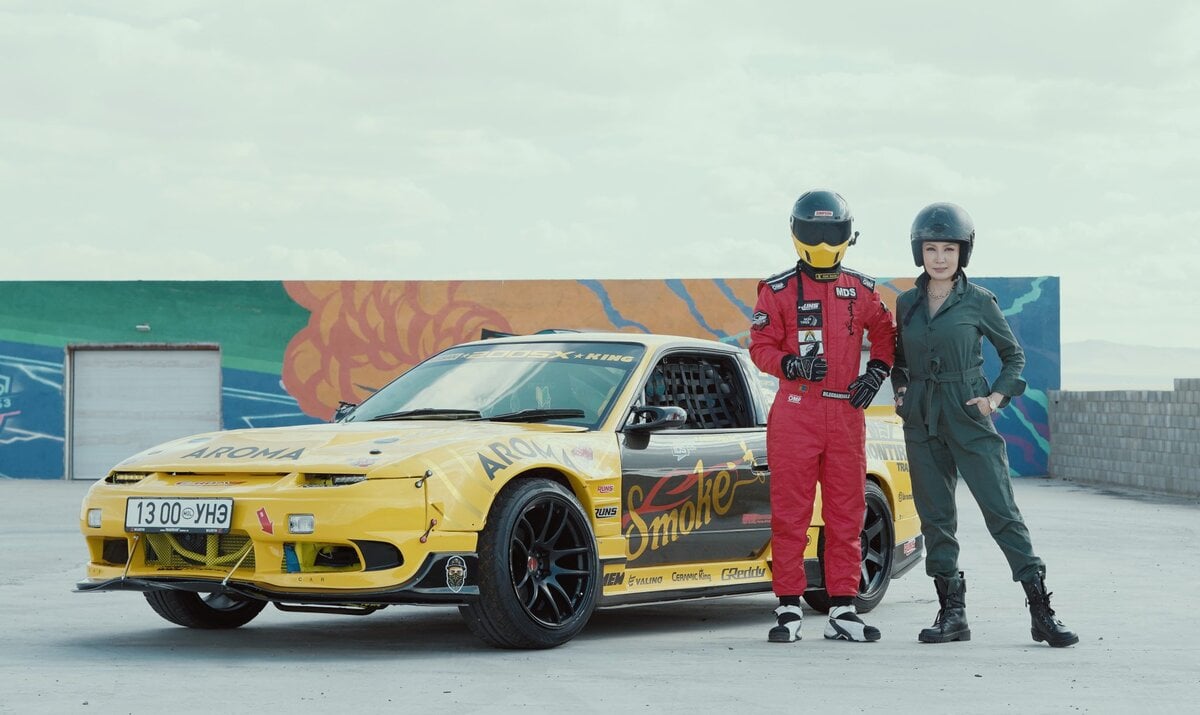 A person in a red racing suit and helmet stands next to a yellow car with graphics. Another person in a military-style jumpsuit and helmet stands beside them. A colorful wall with abstract designs is in the background.