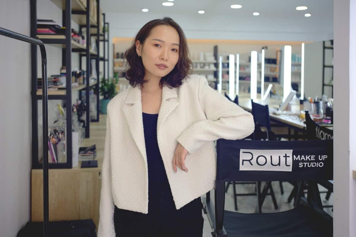 A woman with shoulder-length hair wearing a white jacket stands in a makeup studio. She is leaning against a chair with "Rout Make Up Studio" written on it. Shelves of beauty products are visible in the background.