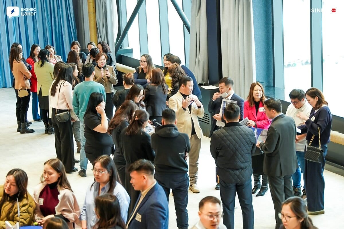 A group of people in a large room engage in conversation. They are dressed in business attire, some standing and others sitting. Large windows provide natural light. The atmosphere appears lively and professional, suggesting a networking event or conference.