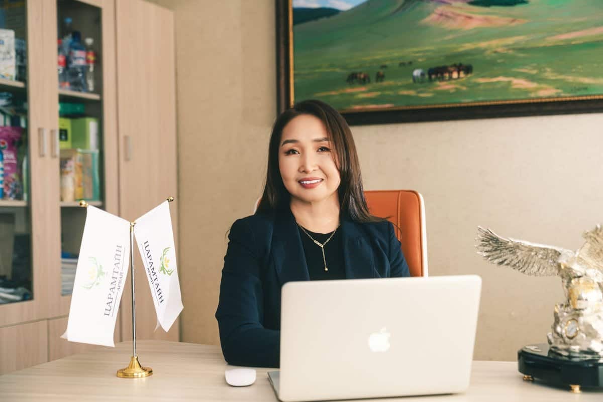 Оффис дээр зөөврийн компьютертэй ширээний ард сууж буй эмэгтэй. Тэр хар хүрэм өмсөж, камер руу инээмсэглэн, хуваарийнхаа хоосон орон зайг хэрхэн дүүргэх талаар бодож байгаа байх. Ширээг хоёр туг чимж, арын ханыг ландшафтын зураг чимдэг. - Business.MN