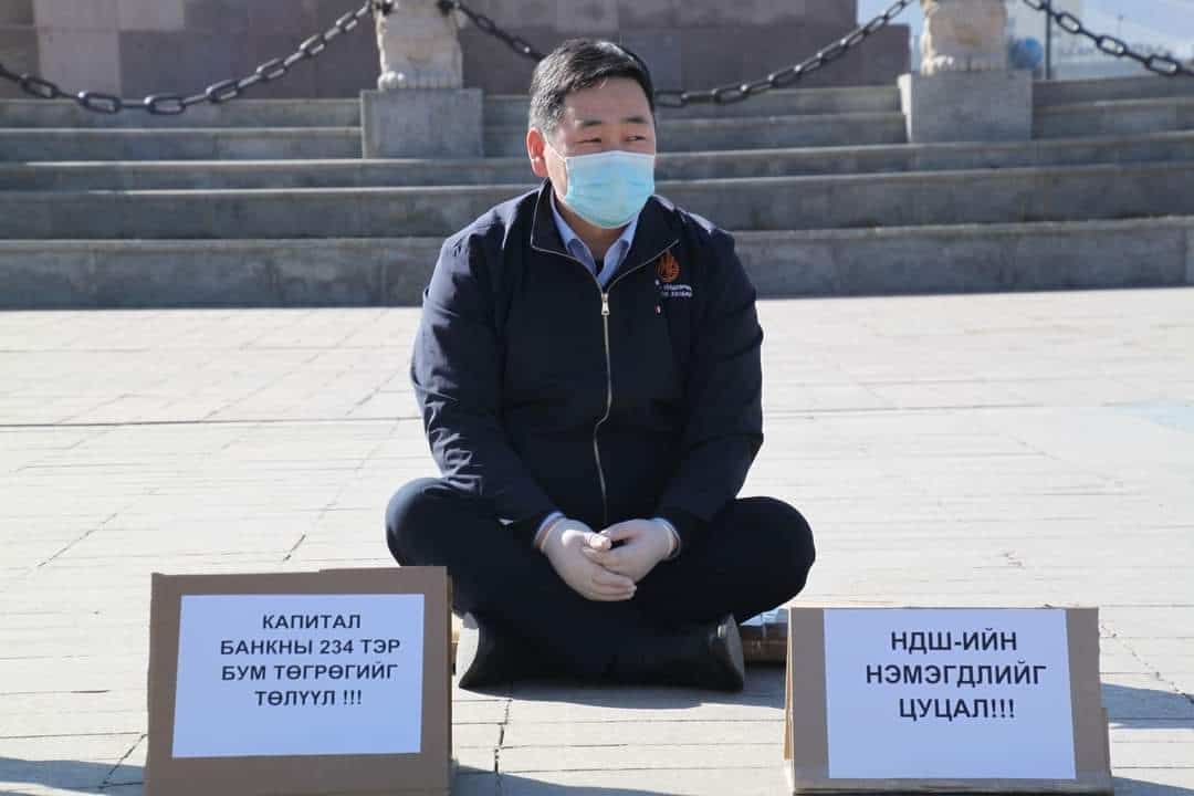 Нүүрний маск зүүж, засмал гадаргуу дээр завилан сууж буй хүн зүүн ази үсгээр бичсэн бичвэр бүхий хоёр цаасан самбар барьж, бизнесээ сурталчилж байна. Арын дэвсгэр дээр чулуун шат, гинжин хаалт орно. - Business.MN