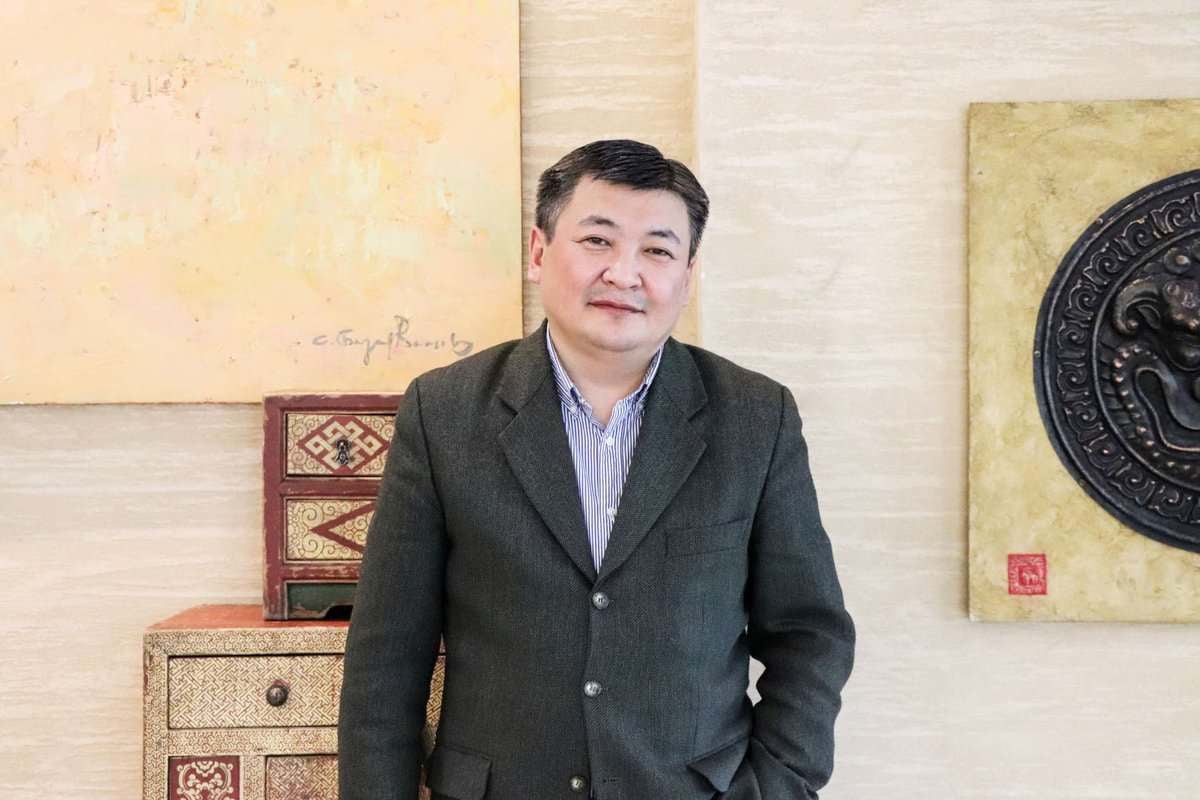 Харанхуй пиджак өмссөн бизнес эрхлэгч шаргал хана, гоёл чимэглэлийн урлаг, модон авдар бүхий өрөөнд зогсож байна. - Business.MN