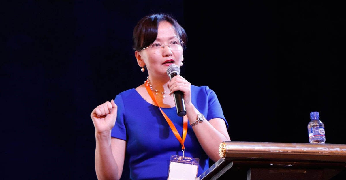 Цэнхэр даашинзтай бизнесмэн эмэгтэй индэр дээр усны савтай зогсож байхдаа микрофоноор ярьж байна. - Business.MN