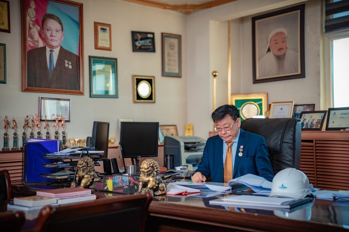 A man in a suit sits at a large desk in an office filled with documents, awards, and framed photos. Two lion statues and a white hard hat are on the desk. Large portraits and certificates decorate the walls.