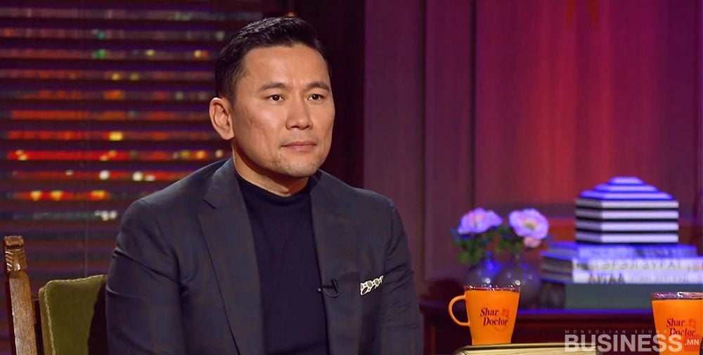 Харанхуй хувцастай хүн дулаан гэрэлтэй өрөөнд сууна. Ойролцоох ширээн дээр "Яригчийн тавцан" гэсэн хоёр улбар шар аяга байна. Цаана нь овоолсон ном, цэцэгтэй жижиг ваар байна. - Business.MN