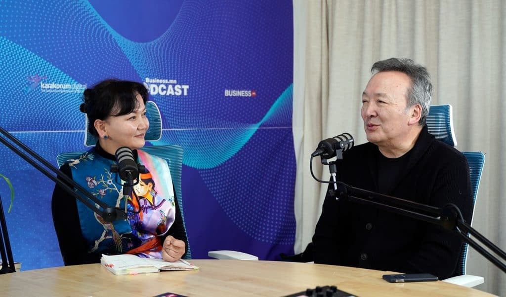 Нэг эмэгтэй, нэг эрэгтэй хоёр хүн микрофонтой ширээний ард суугаад харилцан яриа өрнүүлж байна. Энэ тохиргоо нь подкасттай холбоотой брэндинг агуулсан цэнхэр, ягаан өнгийн дэвсгэр бүхий подкаст студи бололтой. Энтрепренер эмэгтэй инээмсэглэн онгорхой дэвтэр барьдаг. - Business.MN