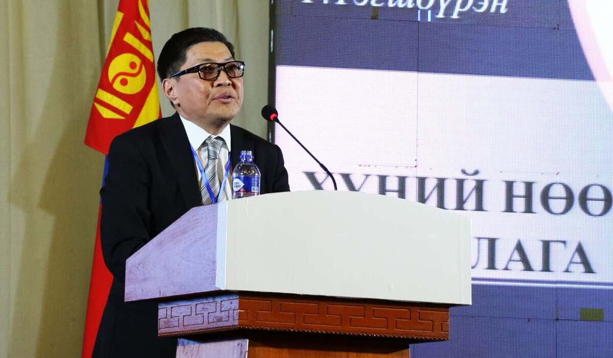 Костюм, зангиа зүүсэн хүн индэр дээр микрофон барьж, энтрепренерийн сүнсийг шингээж, ард нь Монголын далбаатай үг хэлж байна. - Business.MN