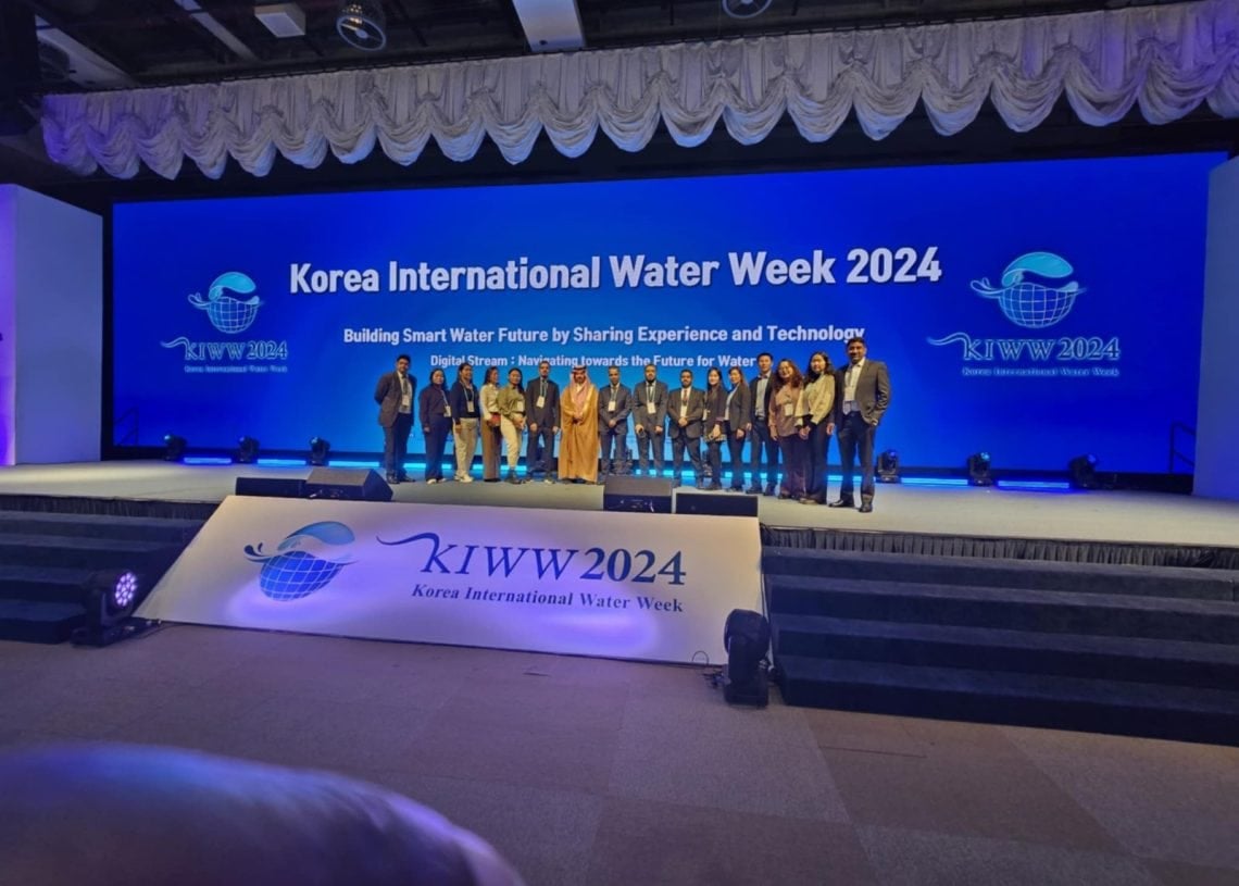 A group of people stands on stage at the Korea International Water Week 2024 event. The backdrop displays the event’s name and slogan, "Building Smart Water Future by Sharing Experience and Technology." Stage lighting creates a vibrant atmosphere.