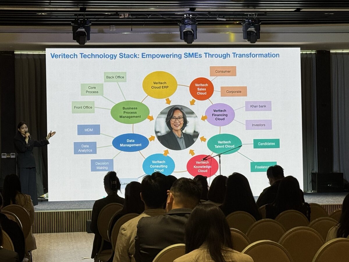 A presenter stands in front of a screen displaying a technology stack diagram for Veritech, with various interconnected business functions, clouds, and data management elements, while an audience sits facing the stage.