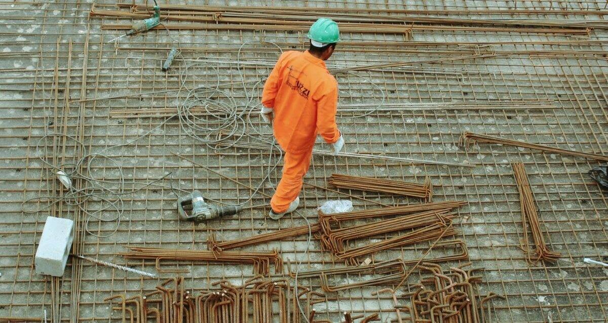 Улбар шар өнгийн комбинезон, ногоон дуулга өмссөн барилгын ажилчин бетонон гадаргуу дээр арматур болон бусад материалуудын дунд зогсож байна. Энтрепренерийн сүнсийг шингээсэн ажилчин талбайн эргэн тойронд тархсан ган арматурын торыг чиглүүлж, цэцэглэн хөгжиж буй бизнест хувь нэмэр оруулдаг. - Business.MN