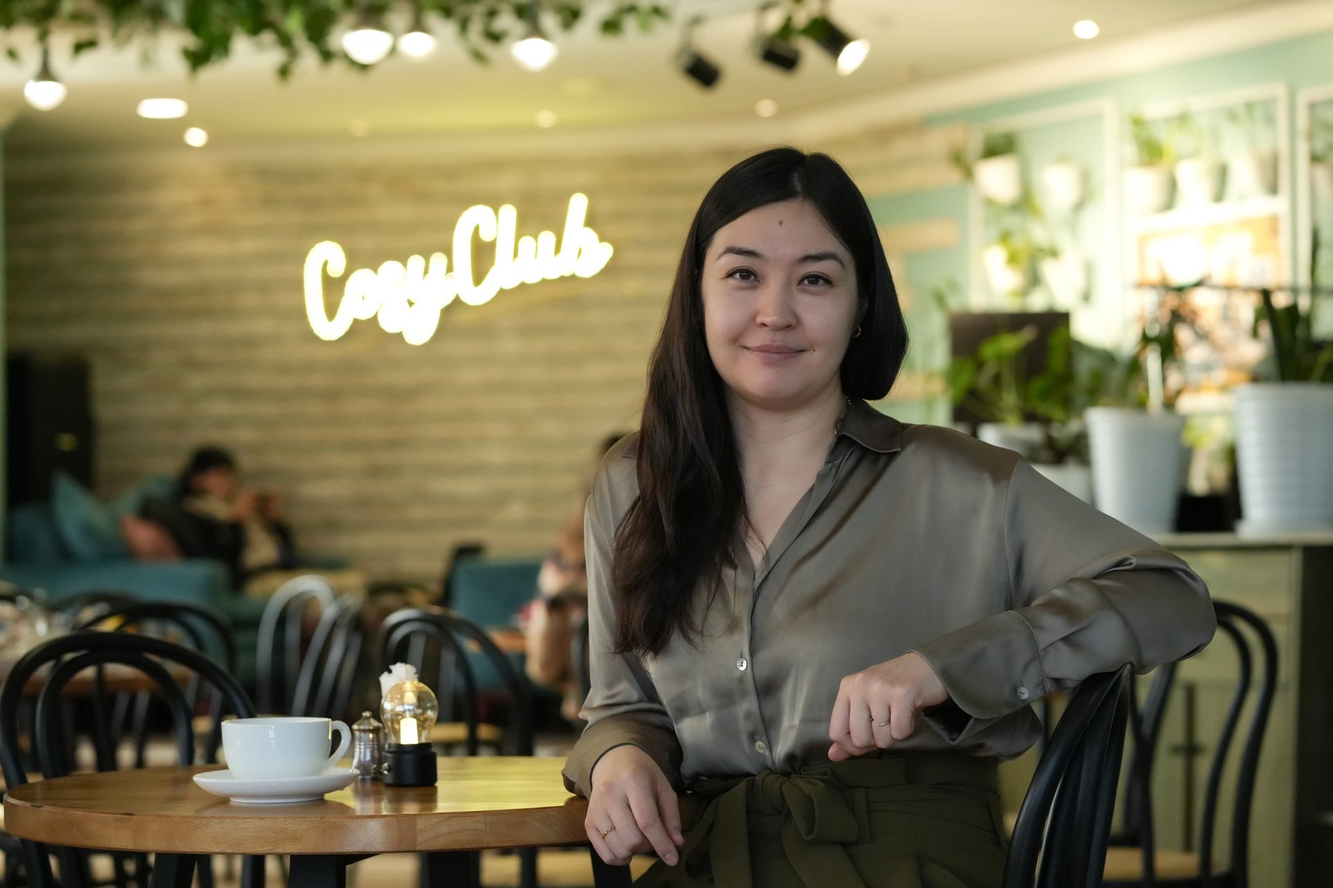 Урт хар үстэй эмэгтэй "Cosy Club" нэртэй тухтай кафед сууж байна. Тэр торгомсог цамц өмсөж, камер руу инээмсэглэж, энтрепренер мэдрэмж төрүүлдэг. Түүний урд ширээн дээр аяга кофе байх бөгөөд арын дэвсгэр нь ургамал, дулаан гэрэлтүүлгээр дүүрэн байдаг. - Business.MN