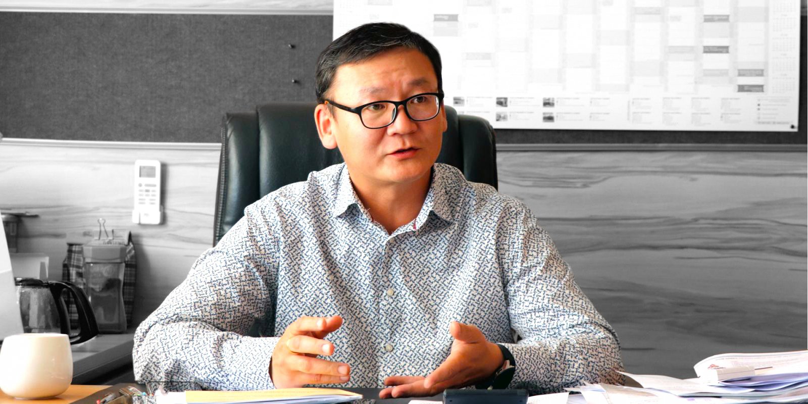 Нүдний шил, хээтэй цамц өмссөн хүн эмх замбараагүй ширээний ард суугаад ярьж, гараараа дохино. Бүдгэрсэн дэвсгэр дээр үйсэн самбар болон хуанли харагдана. - Business.MN