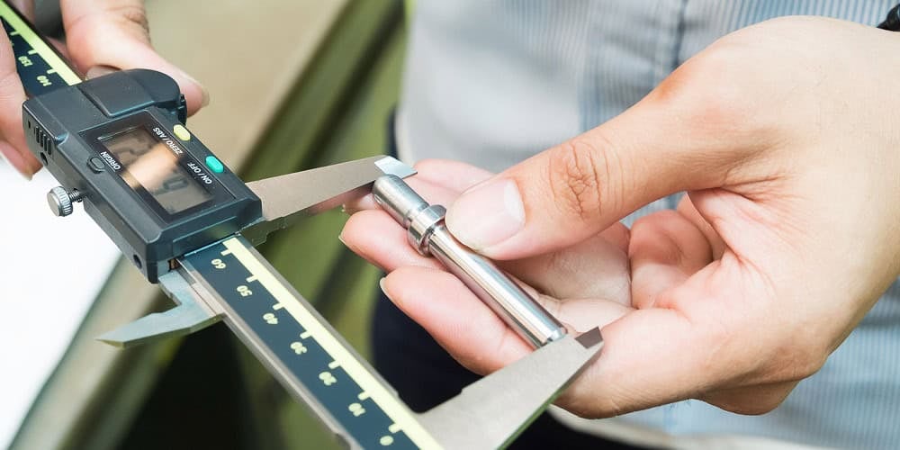A person using a digital caliper to measure a small metal cylindrical object demonstrates чанараар манлайлах. The caliper displays measurements on its digital screen, while the person holds the object in their other hand, emphasizing precision. The background is blurred.