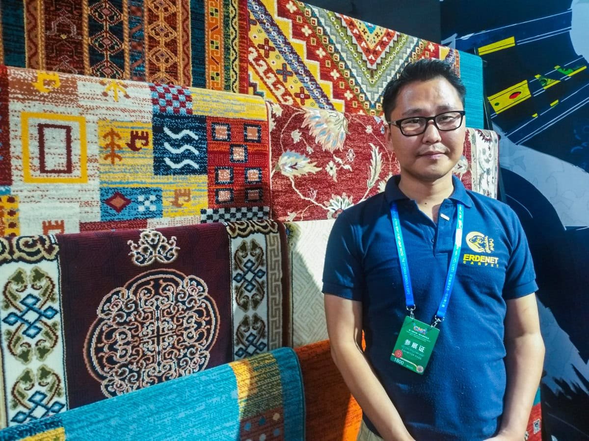 Нүдний шил, цэнхэр поло цамц өмссөн эрэгтэй өнгөлөг, хээтэй хивсний үзүүлэнгийн өмнө зогсож байна. Тэрээр хүзүүндээ үйл явдлын тэмдэгтэй байдаг. Арын дэвсгэр нь янз бүрийн нарийн төвөгтэй дизайн, бүтэцтэй. - Business.MN