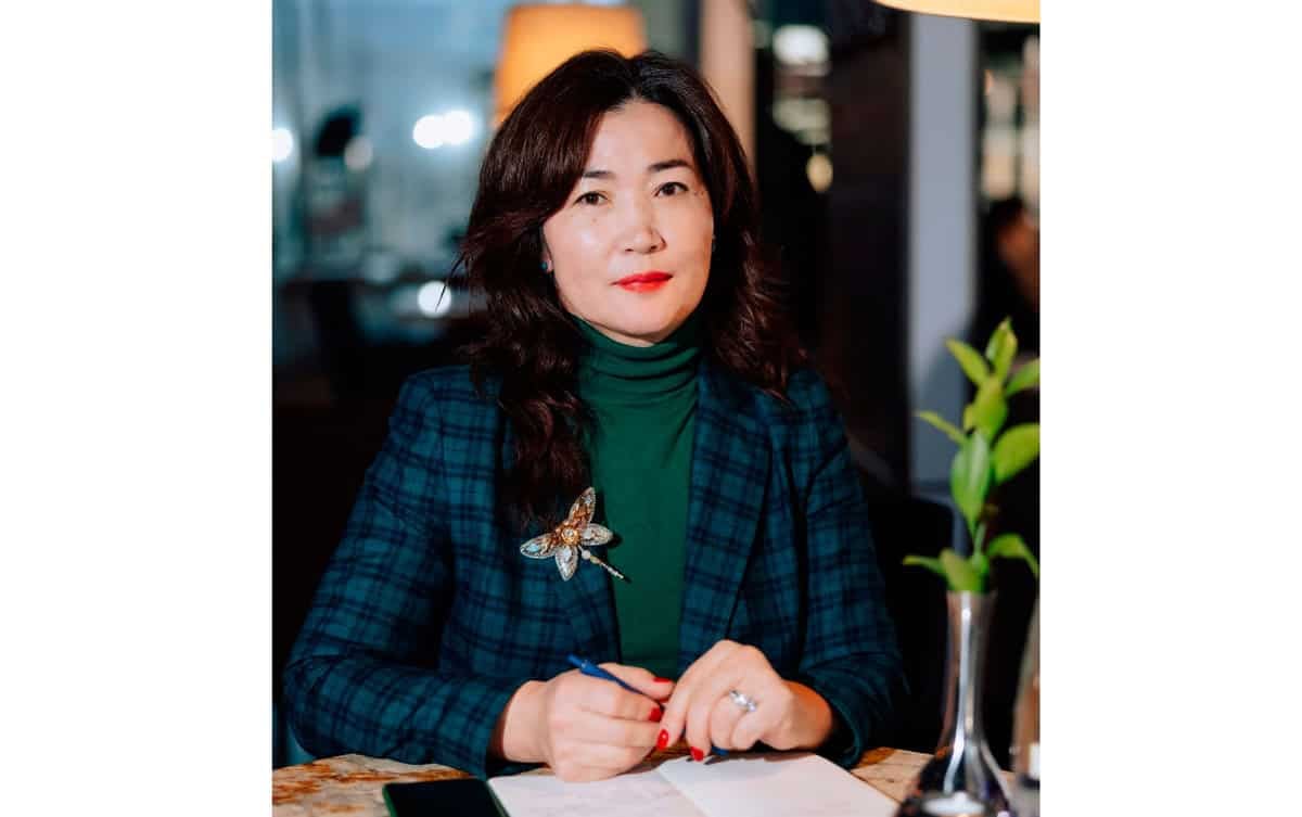 Ширээн дээр ногоон, цэнхэр алаг пиджак өмссөн, урт долгионтой үстэй эмэгтэй сууж байна. Тэр үзэг барьдаг бөгөөд урд нь бичиг баримтууд байдаг нь түүний бизнестэй холбоотой байх магадлалтай. Цаана нь дэнлүү, вааранд хийсэн жижиг ногоон ургамал харагдана. Тэр эрвээхэй энгэрийн зүүсэн байна. - Business.MN