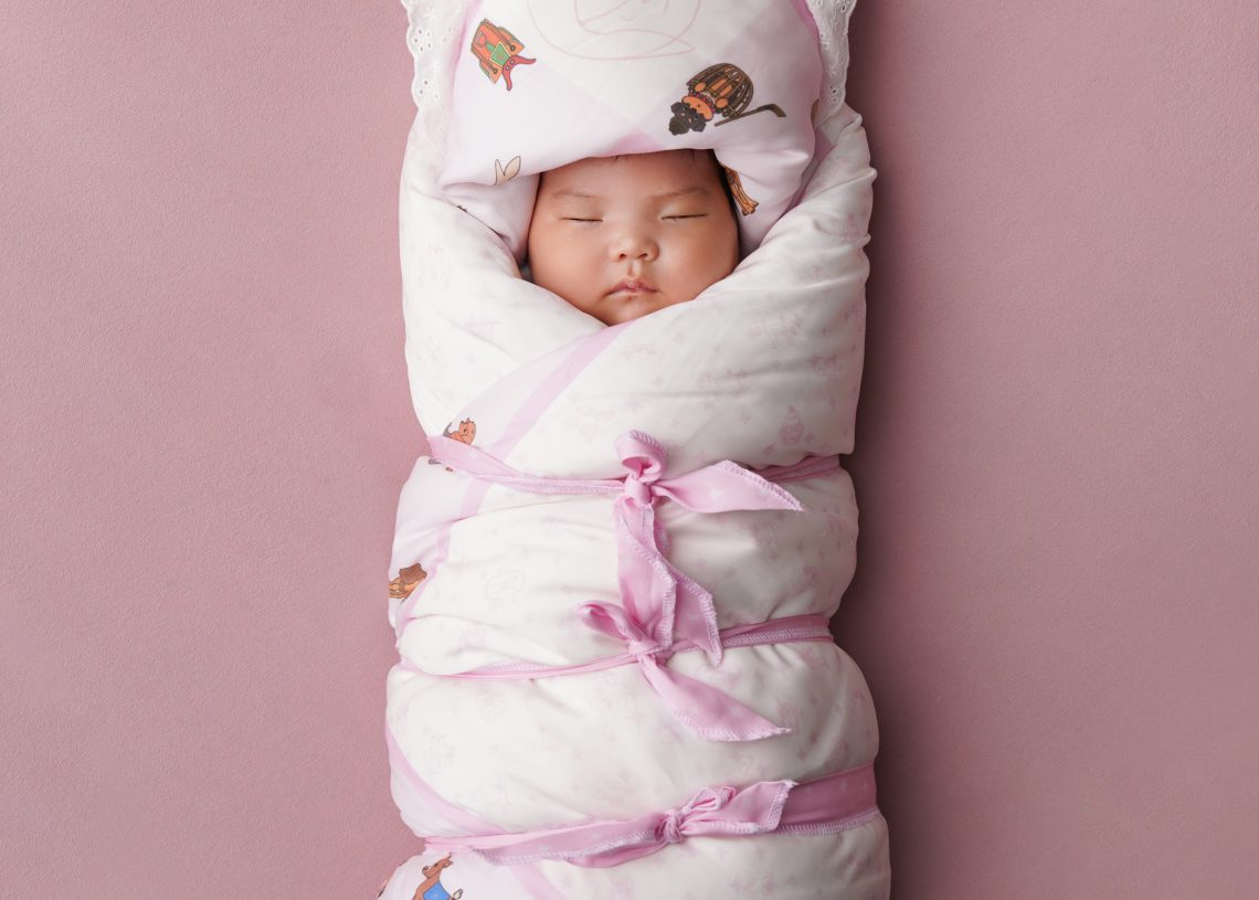 Нярай хүүхэд амьтдын дүрс бүхий цагаан, ягаан хээтэй хөнжлөөр сайтар ороож, зөөлөн ягаан гадаргуу дээр хэвтэж, тайван унтаж байна. - Business.MN