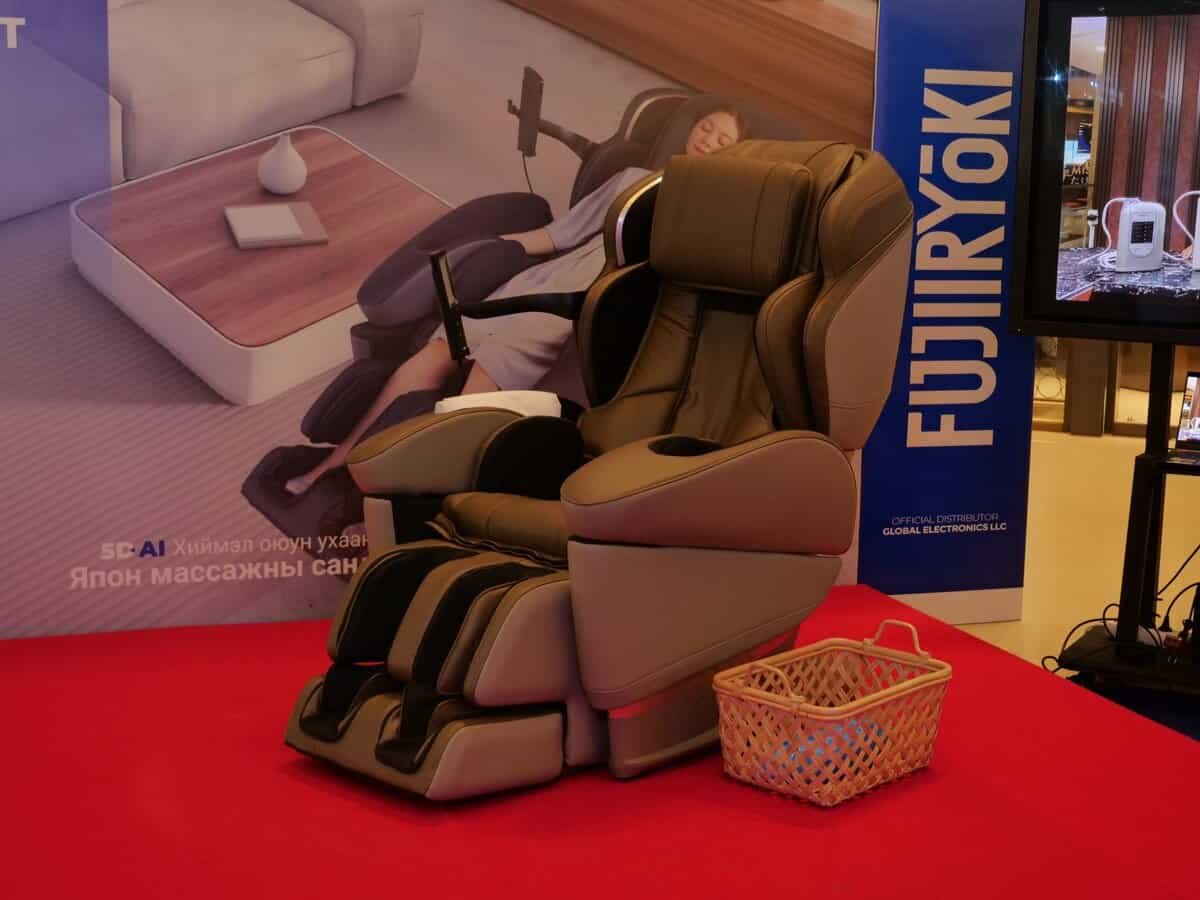 Саарал, хар бүрээстэй өндөр технологийн массажны сандал улаан хивсэн дээр тавигдсан нь энтрепренерийн сүнсийг илэрхийлдэг. Зэгсэн сагс сандлын дэргэд сууна. Ард байрлах том зурагт хуудас нь брендинг "FUJIIRYOKI" болон зочны өрөөний загварлаг зохион байгуулалтын зургуудтай. - Business.MN